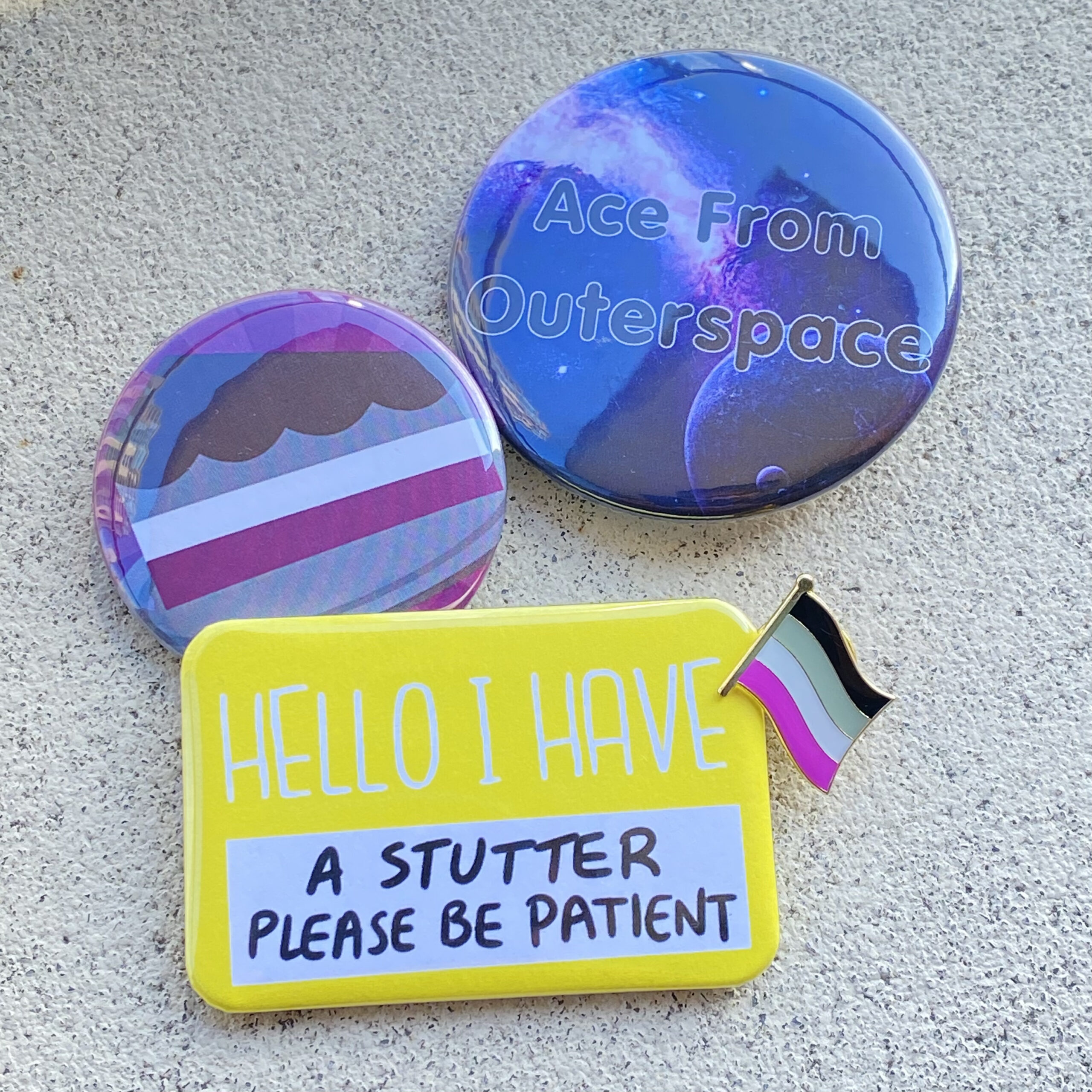 Four buttons (the kind pinned to a shirt or bag) on a cement floor: One says "Ace from Outer Space", one is a picture of a cake in the asexual pride flag colours, and one is a yellow rectangle that says, "Hello, I have a stutter, please be patient". At the upper right-hand corner of the yellow button is a small pin of the asexual pride flag.